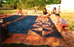 Painting the Tapa Cloth: The tapa cloth is laid out on the grass for painting. In this photograph members of the Vaka family from Lapaha are painting it with black dye made from the bark and roots of the koka tree. It is a slow and difficult process to make the dye.